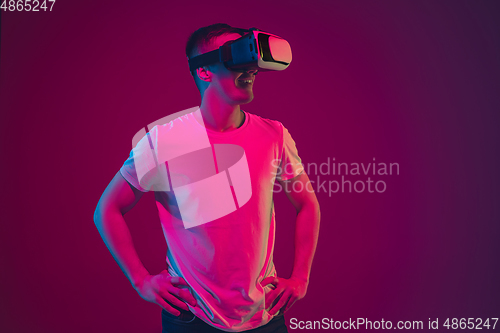 Image of Caucasian man\'s portrait isolated on pink-purple studio background in neon light, playing with VR-headset