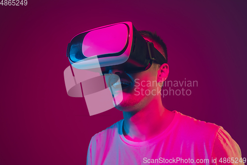 Image of Caucasian man\'s portrait isolated on pink-purple studio background in neon light, playing with VR-headset