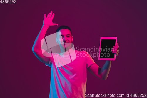 Image of Caucasian man\'s portrait isolated on pink-purple studio background in neon light, using tablet