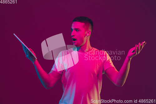 Image of Caucasian man\'s portrait isolated on pink-purple studio background in neon light, using tablet