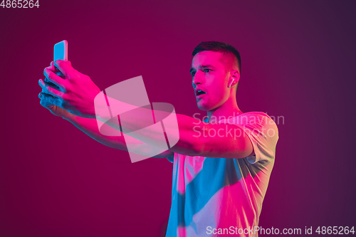 Image of Caucasian man\'s portrait isolated on pink-purple studio background in neon light, with smartphone and earphones