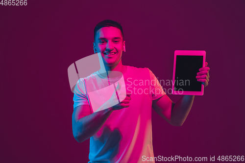 Image of Caucasian man\'s portrait isolated on pink-purple studio background in neon light, using tablet