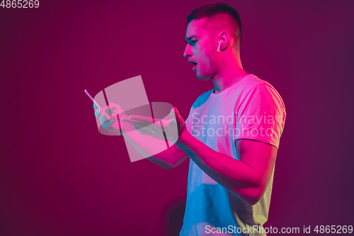 Image of Caucasian man\'s portrait isolated on pink-purple studio background in neon light, with smartphone and earphones