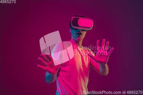 Image of Caucasian man\'s portrait isolated on pink-purple studio background in neon light, playing with VR-headset