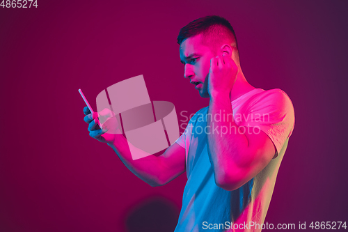 Image of Caucasian man\'s portrait isolated on pink-purple studio background in neon light, with smartphone and earphones