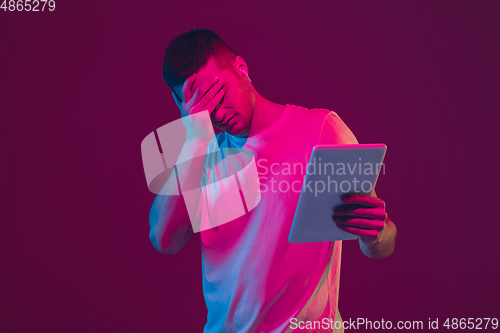 Image of Caucasian man\'s portrait isolated on pink-purple studio background in neon light, using tablet