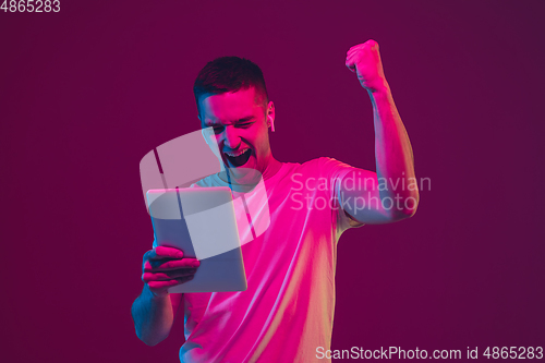 Image of Caucasian man\'s portrait isolated on pink-purple studio background in neon light, using tablet