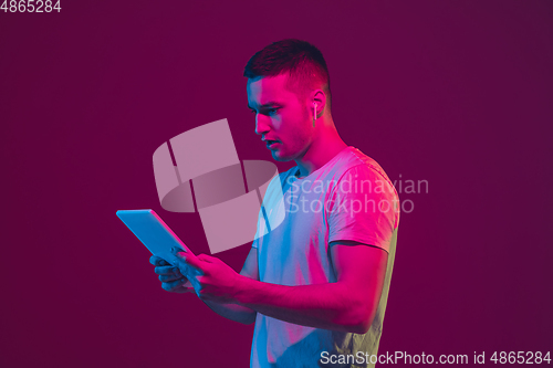 Image of Caucasian man\'s portrait isolated on pink-purple studio background in neon light, using tablet
