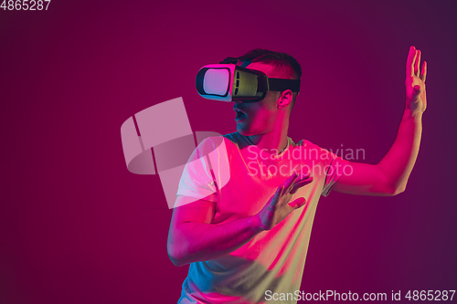 Image of Caucasian man\'s portrait isolated on pink-purple studio background in neon light, playing with VR-headset