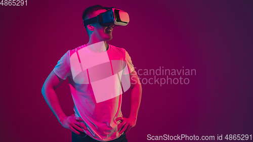 Image of Caucasian man\'s portrait isolated on pink-purple studio background in neon light, playing with VR-headset