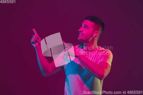 Image of Caucasian man\'s portrait isolated on pink-purple studio background in neon light, using wireless earphones
