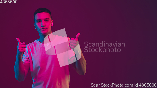 Image of Caucasian man\'s portrait isolated on pink-purple studio background in neon light, using wireless earphones