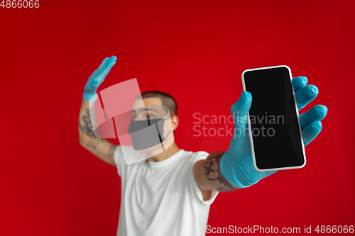 Image of Caucasian young man in protective face mask and medical gloves on red studio background - showing blank phone\'s screen