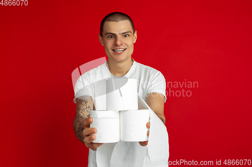 Image of Caucasian young man\'s portrait on red studio background - holding toilet papers, essential goods during quarantine and self-insulation