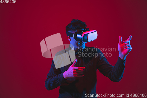 Image of Caucasian man\'s portrait isolated on red studio background in purple-blue neon light, emotional and beautiful, playing wore VR-headset