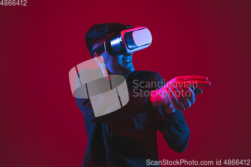 Image of Caucasian man\'s portrait isolated on red studio background in purple-blue neon light, emotional and beautiful, playing wore VR-headset