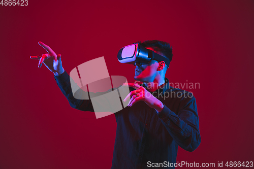 Image of Caucasian man\'s portrait isolated on red studio background in purple-blue neon light, emotional and beautiful, playing wore VR-headset