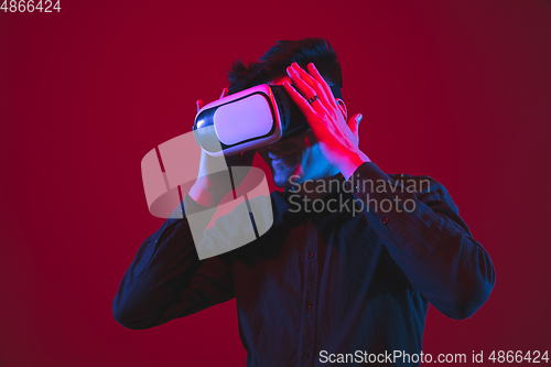 Image of Caucasian man\'s portrait isolated on red studio background in purple-blue neon light, emotional and beautiful, playing wore VR-headset