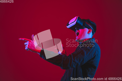 Image of Caucasian man\'s portrait isolated on red studio background in purple-blue neon light, emotional and beautiful, playing wore VR-headset