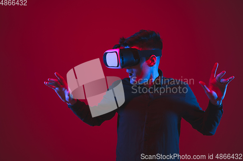 Image of Caucasian man\'s portrait isolated on red studio background in purple-blue neon light, emotional and beautiful, playing wore VR-headset