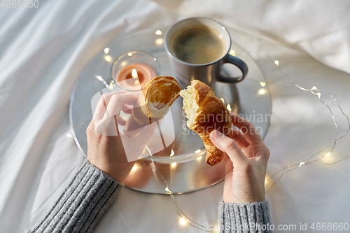 Image of hands of woman eating croissant with coffee in bed