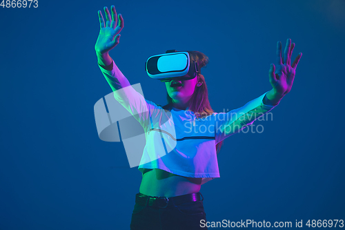 Image of Caucasian woman\'s portrait isolated on blue studio background in pink-green neon light, stylish and beautiful, playing with VR-headset