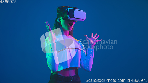 Image of Caucasian woman\'s portrait isolated on blue studio background in pink-green neon light, stylish and beautiful, playing with VR-headset