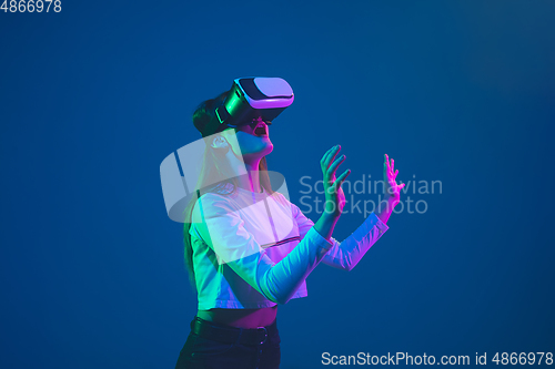 Image of Caucasian woman\'s portrait isolated on blue studio background in pink-green neon light, stylish and beautiful, playing with VR-headset