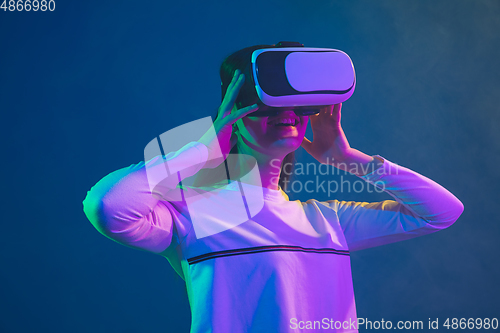 Image of Caucasian woman\'s portrait isolated on blue studio background in pink-green neon light, stylish and beautiful, playing with VR-headset