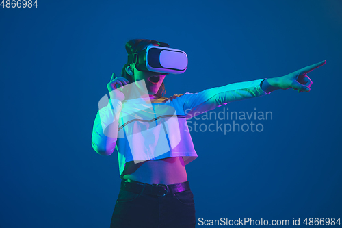 Image of Caucasian woman\'s portrait isolated on blue studio background in pink-green neon light, stylish and beautiful, playing with VR-headset