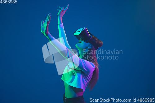 Image of Caucasian woman\'s portrait isolated on blue studio background in pink-green neon light, stylish and beautiful, playing with VR-headset