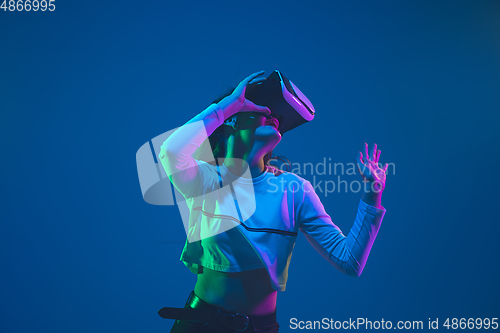 Image of Caucasian woman\'s portrait isolated on blue studio background in pink-green neon light, stylish and beautiful, playing with VR-headset