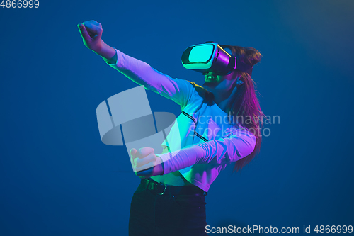 Image of Caucasian woman\'s portrait isolated on blue studio background in pink-green neon light, stylish and beautiful, playing with VR-headset