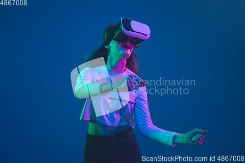 Image of Caucasian woman\'s portrait isolated on blue studio background in pink-green neon light, stylish and beautiful, playing with VR-headset