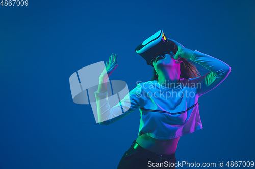Image of Caucasian woman\'s portrait isolated on blue studio background in pink-green neon light, stylish and beautiful, playing with VR-headset