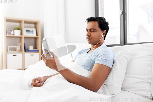 Image of indian man in earphones with phone in bed at home