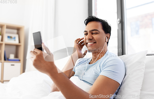 Image of indian man in earphones with phone in bed at home