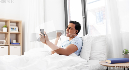 Image of happy indian man with smartphone in bed at home