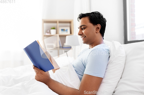 Image of happy indian man reading book in bed at home