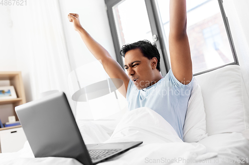 Image of happy indian man with laptop in bed at home