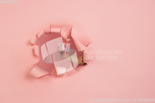 Image of Female hand giving gift in torn coral, pink paper hole background, celebration