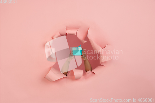 Image of Female hand giving gift in torn coral, pink paper hole background, celebration