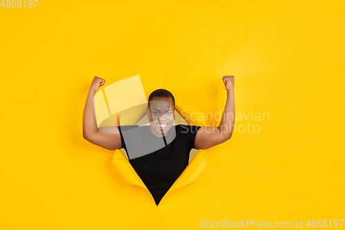Image of Cheerful young woman poses in torn yellow paper hole background, emotional and expressive