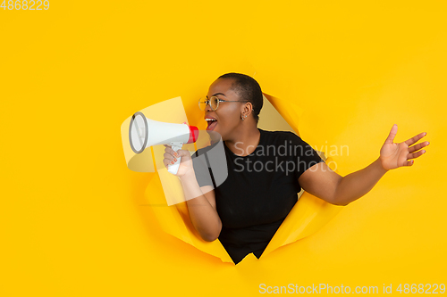 Image of Cheerful young woman poses in torn yellow paper hole background, emotional and expressive, shouting and calling with speaker