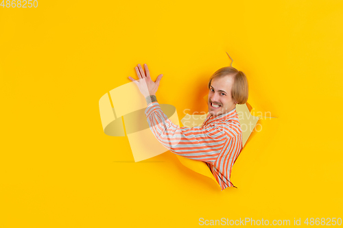 Image of Cheerful young man poses in torn yellow paper hole background, emotional and expressive