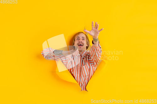 Image of Cheerful young man poses in torn yellow paper hole background, emotional and expressive