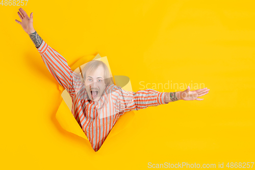 Image of Cheerful young man poses in torn yellow paper hole background, emotional and expressive