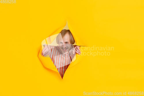 Image of Cheerful young man poses in torn yellow paper hole background, emotional and expressive