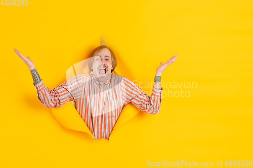 Image of Cheerful young man poses in torn yellow paper hole background, emotional and expressive