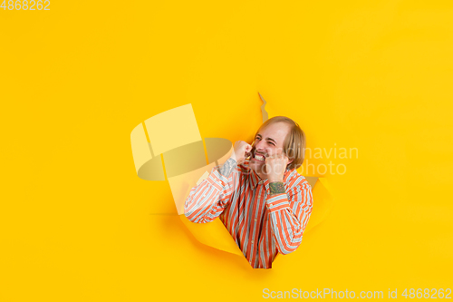 Image of Cheerful young man poses in torn yellow paper hole background, emotional and expressive